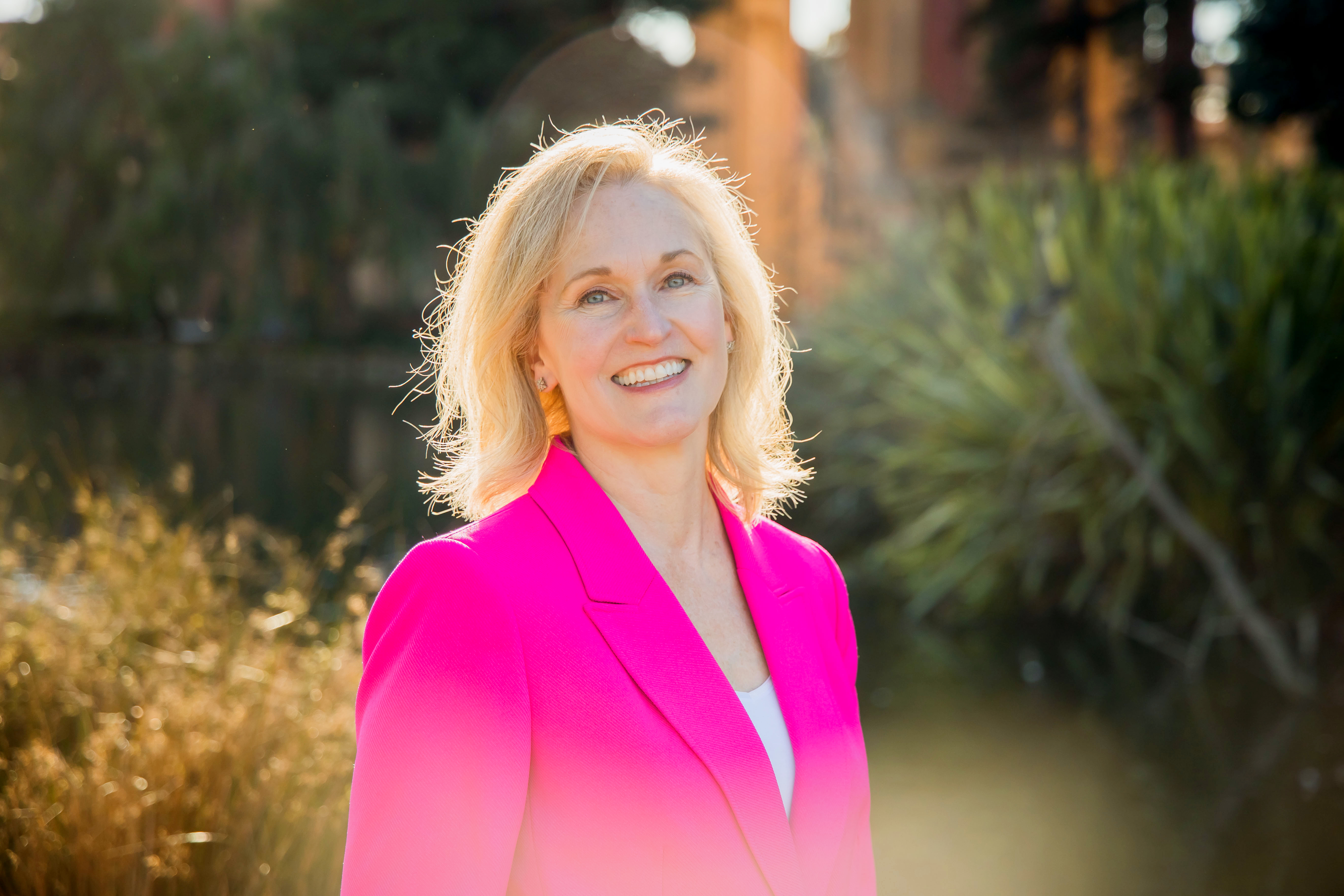 Susan M. Davis with blonder hair poses outdoors wearing a fuschia blazer