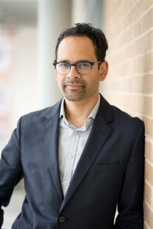 Sunil Amrith poses against a brick wall wearing a navy blazer and light blue shirt