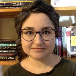 Person with short hair and glasses, standing in front of a bookshelf filled with books.
