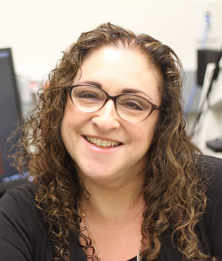 A smiling person wearing glasses and with curly hair, seated indoors in an office setting.