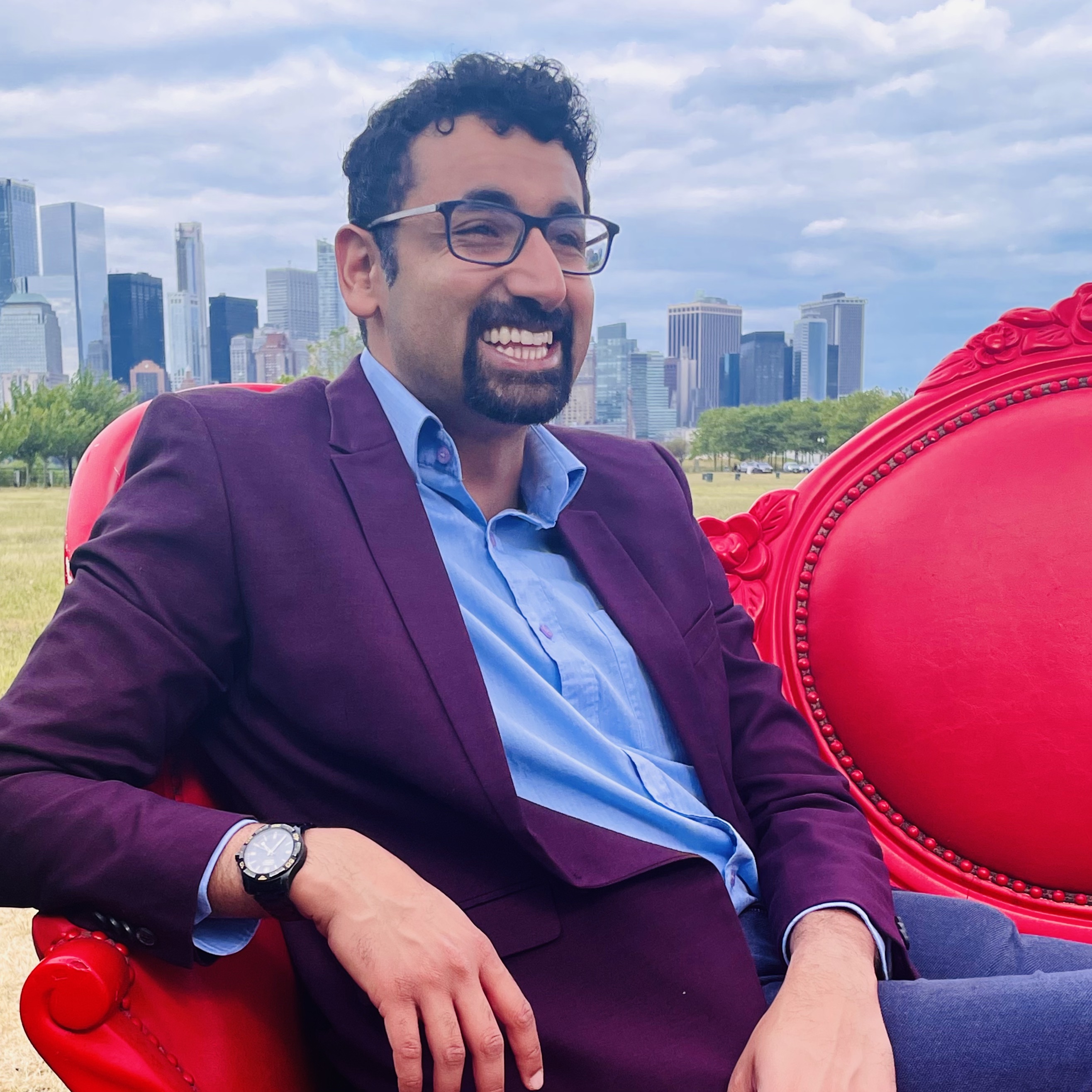 Person in a maroon blazer and blue shirt, laughing while seated on a red armchair in a park with the Chicago skyline in the background.