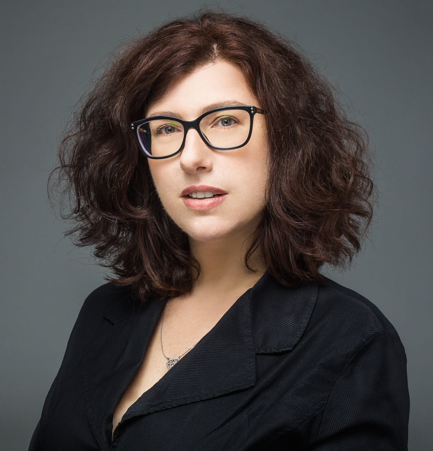 Portrait of a person wearing glasses and a black blazer against a grey background.