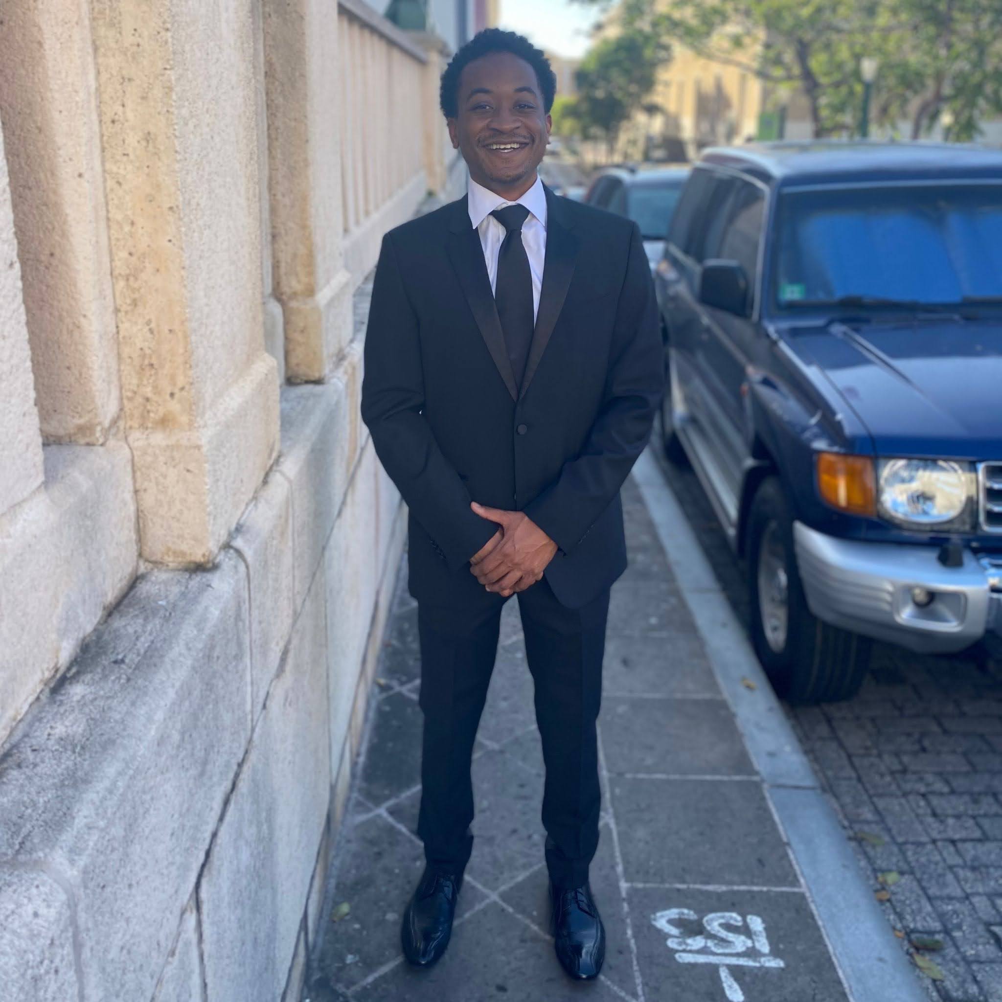 Person in a black suit smiling while standing on a sidewalk next to a stone wall.