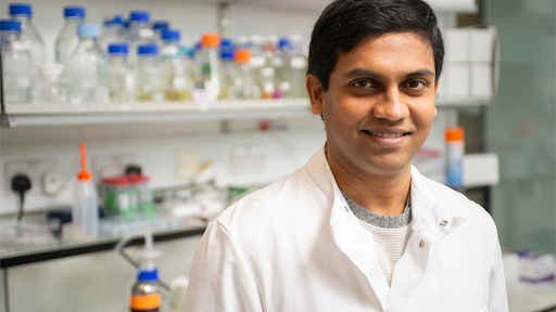 Scientist in a lab coat smiling at the camera in a laboratory filled with chemical bottles and lab equipment.