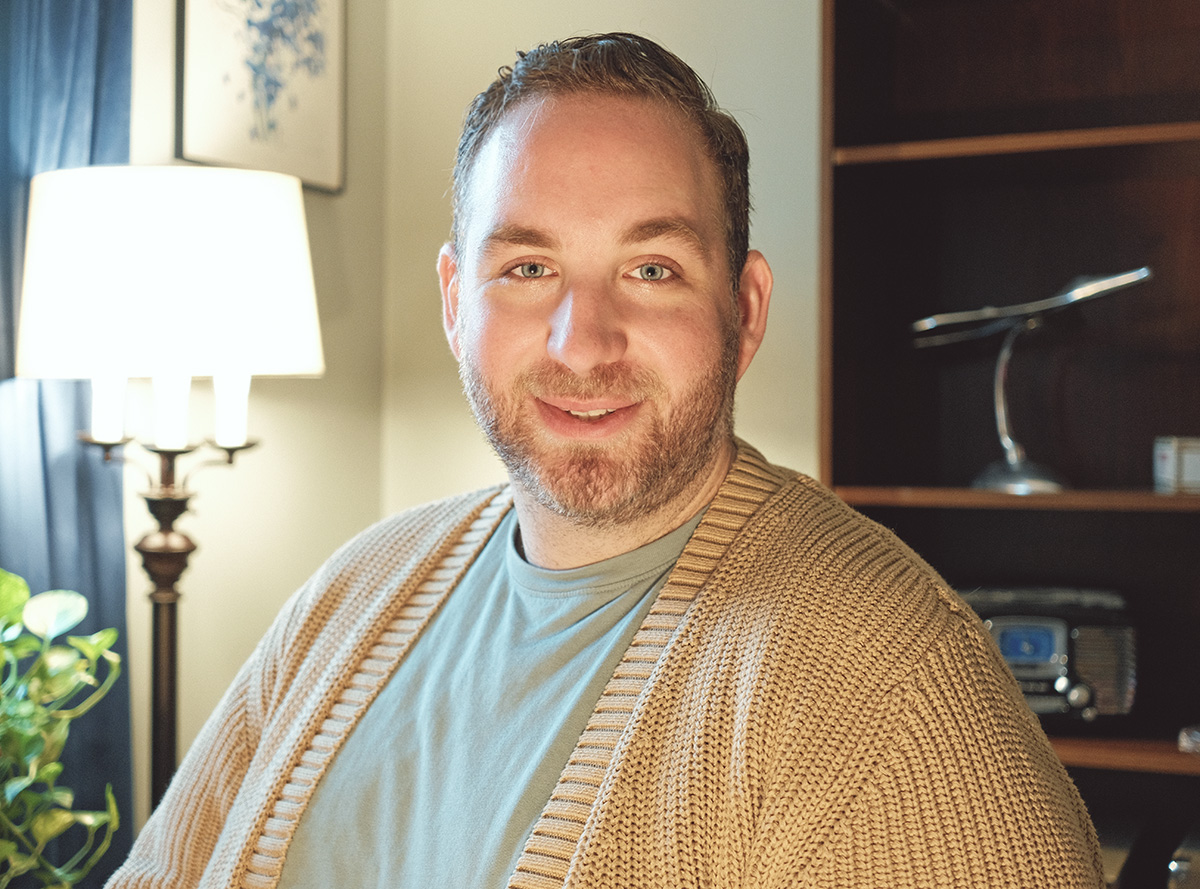 Portrait of an individual smiling, seated in a cozy home office with elegant decor, including a green plant and a vintage desk lamp.