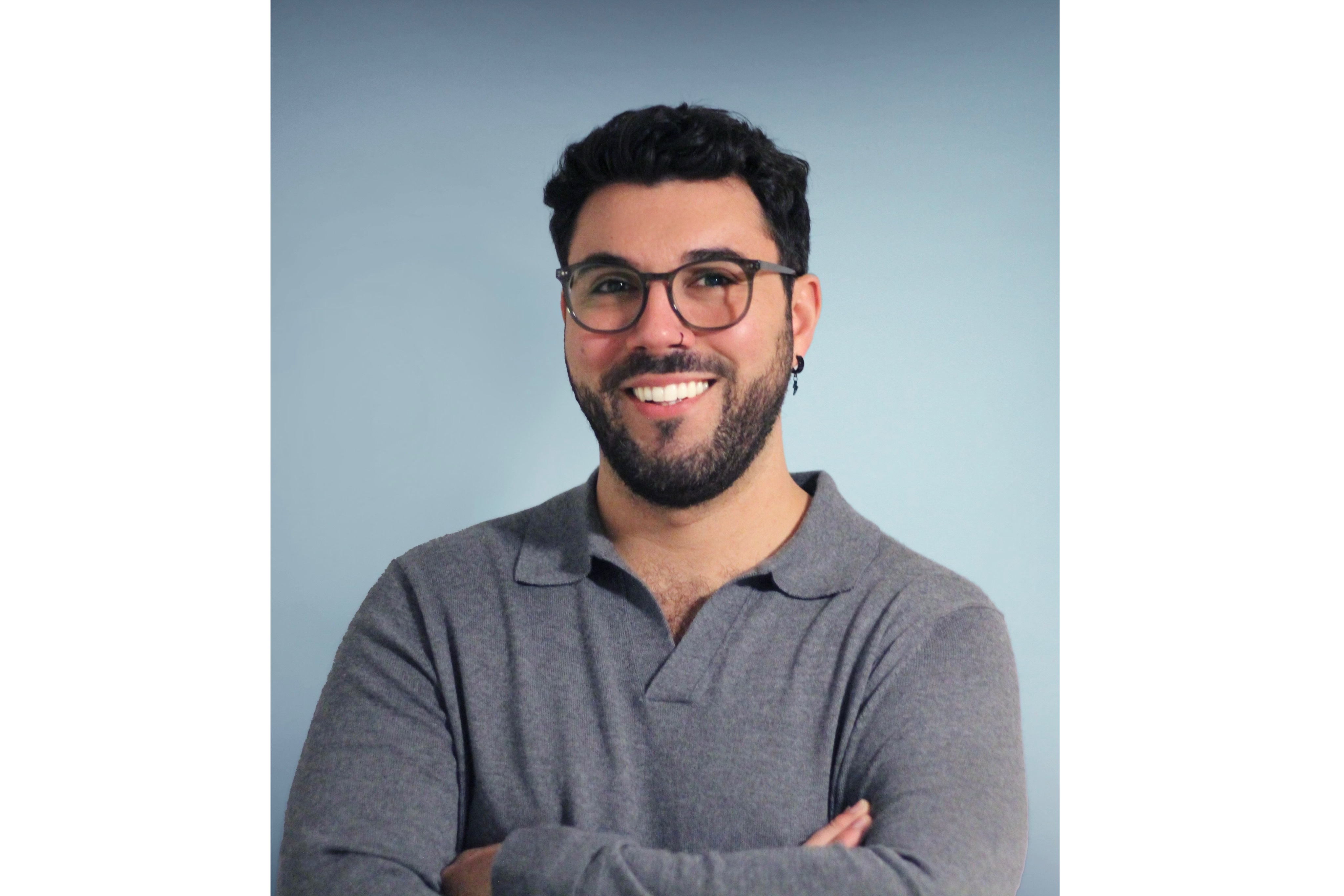 A smiling man with glasses and a beard stands against a light blue background. He is wearing a gray, long-sleeved, collared shirt and has his arms crossed.
