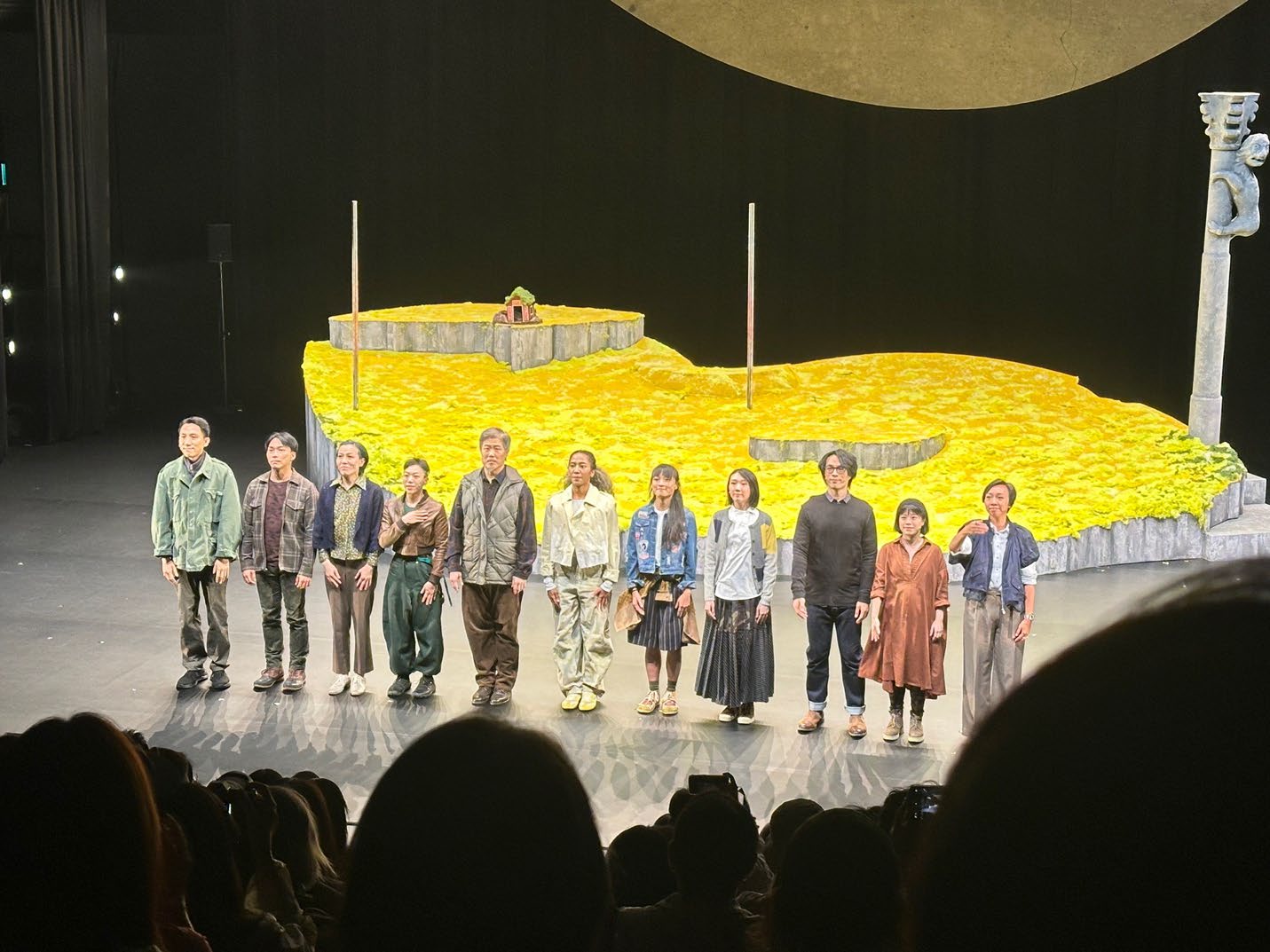 Cast members of a theater production taking a bow on stage, featuring a set designed to look like a golden field under a large circular backdrop.