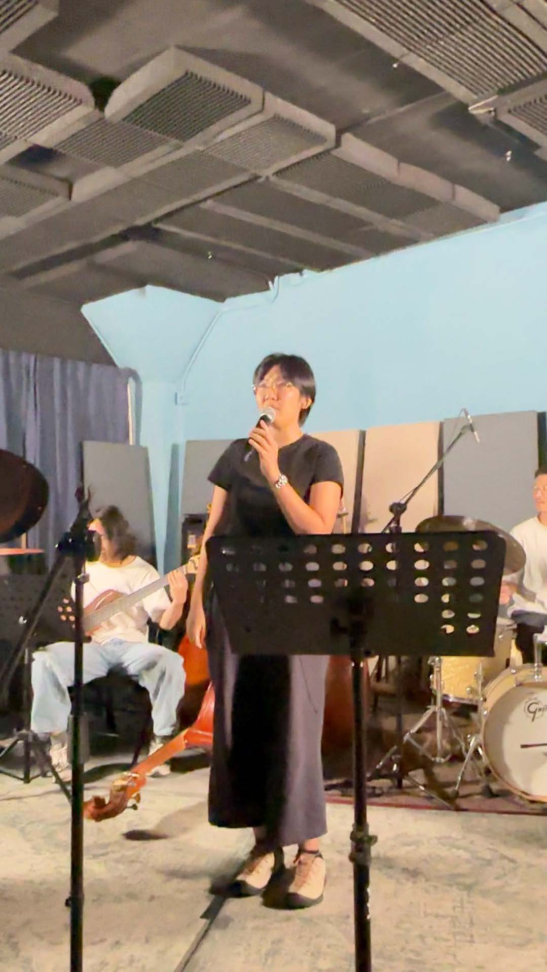 Person singing into a microphone with a band performing in the background in a room with overhead ductwork.