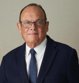 Individual wearing glasses and a dark suit poses in front of a tan background