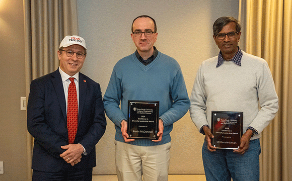 Three men stand together indoors, smiling. The two on the right hold plaques. They're dressed in business casual attire, conveying a celebratory mood.