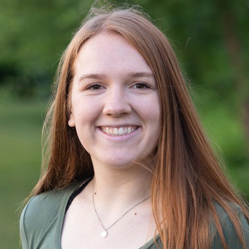 A headshot of a smiling young woman with long, straight strawberry-blonde hair. She is wearing a green top and a small silver pendant necklace. The background consists of vibrant, out-of-focus green foliage.