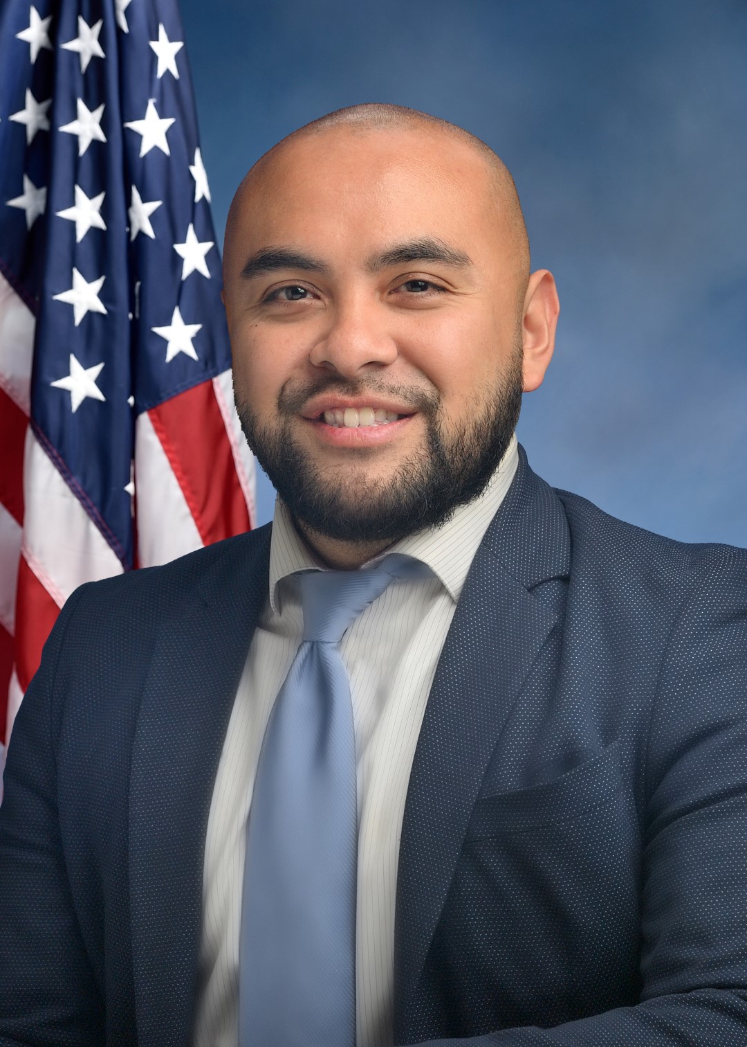 Steven Raga A professional headshot of a smiling man with a shaved head and a dark beard. He is wearing a blue patterned blazer, a light-colored pinstriped shirt, and a solid light-blue tie. An American flag is visible in the background to the left against a blue studio backdrop.